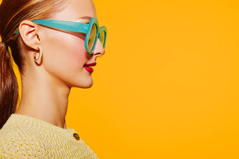 Profile of a young woman wearing large turquoise glasses and a yellow sweater against a bright orange background. Concept of fashion, style, and vibrant colors.