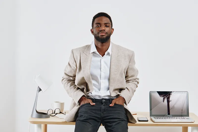 Confident young man with dark skin, stylish beige blazer, white shirt, and jeans sitting on desk with modern workspace setup, minimalistic background, and natural lighting. Business casual fashion, pr