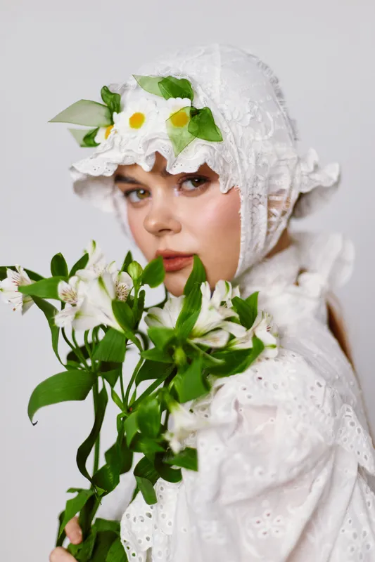 Young woman in a floral headpiece holding white flowers, wearing a delicate white dress, expressing serenity and nature s beauty in a bright, soft toned setting
