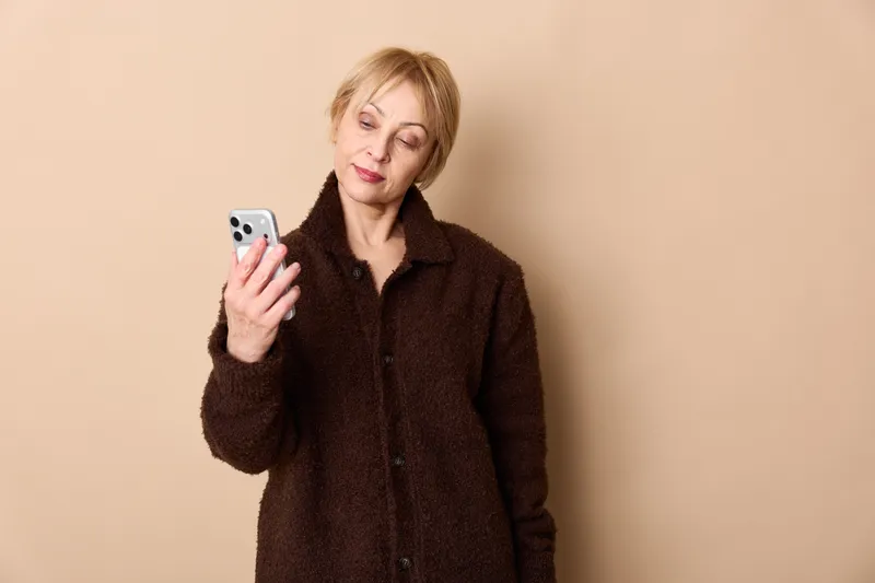 Woman smartphone in studio portrait, older technology anxiety and loneliness shown by concerned expression, she holds phone indoors against soft beige background
