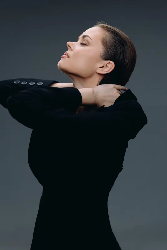 Elegant woman in black outfit posing with closed eyes and serene expression on dark background.