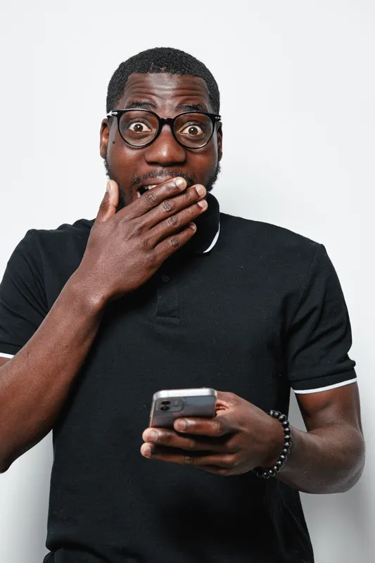 African American man with dark skin and glasses holding smartphone with surprised expression, wearing black polo shirt on white background in studio portrait.