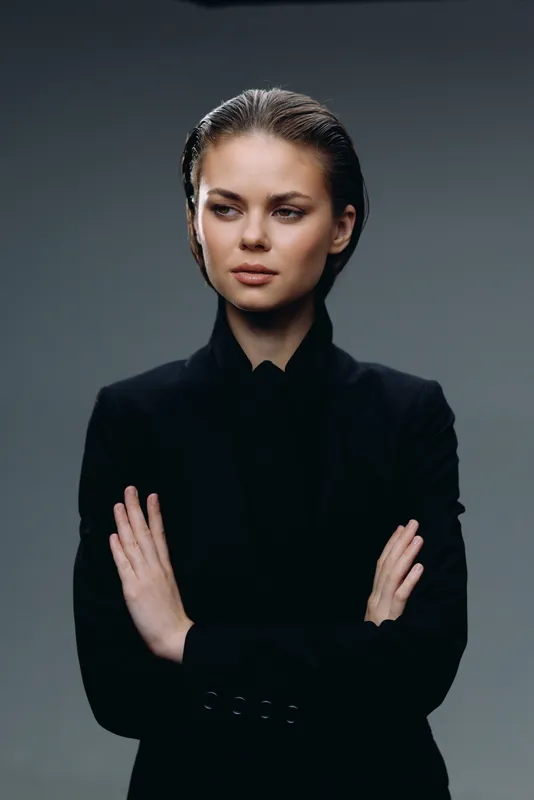 Serious woman in black blazer with crossed arms on gray background expressing confidence and strength.