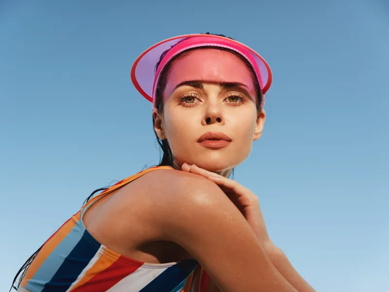 Portrait of a confident woman in a striped swimsuit wearing a pink visor, posed with a graceful arm, against a clear blue sky and warm sunlight.