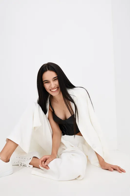 Smiling woman with long black hair in white pants and jacket sitting on floor, wearing black top, expressing happiness, casual fashion in bright studio setting.