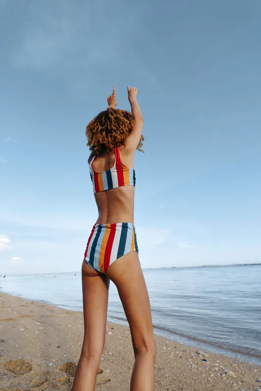 Young woman with curly hair wearing a colorful striped swimsuit raises her arms joyfully while standing on a sandy beach near calm water under a clear blue sky.