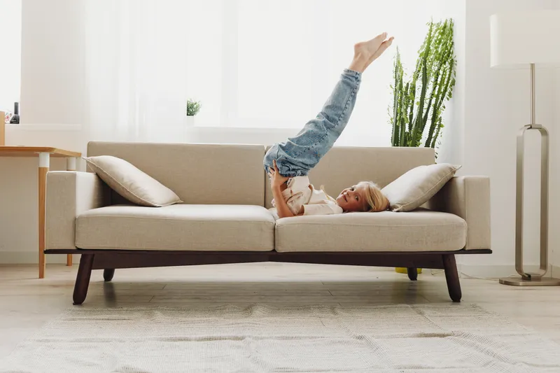 Young boy in casual clothes lying upside down on beige sofa in bright living room. Cozy home interior with plants and soft natural light creates relaxed atmosphere for leisure.