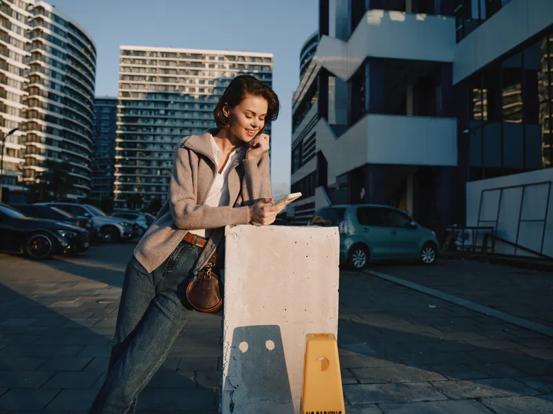 A professional woman stands outdoors in a modern city setting, using a smartphone with a smile, exuding confidence and focus as she navigates a busy day and communicates with ease in a vibrant urban