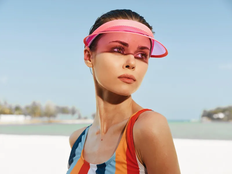 Portrait of a young woman on a sunny beach, wearing a pink visor and a colorful striped swimsuit, gazing into the distance with confident, serene poise.