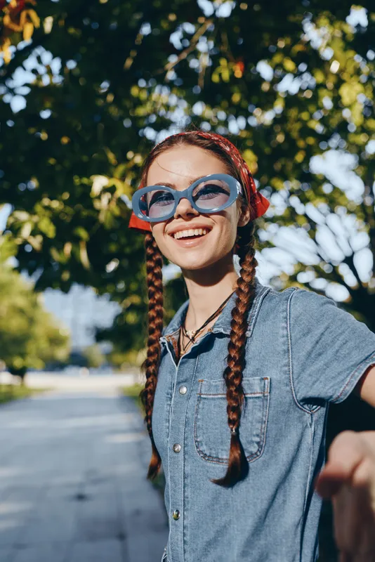 lifestyle woman in modern boho-western denim street style wearing blue sunglasses and red bandana, smiling outdoors with natural green background, film color tones