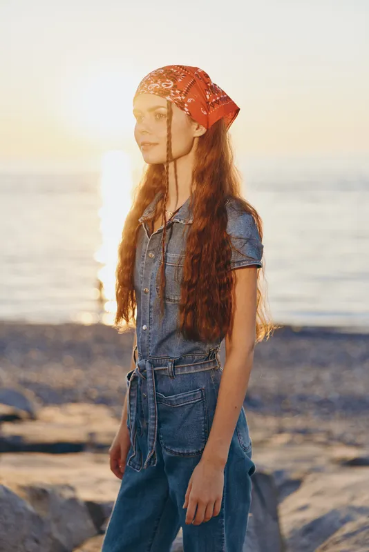 lifestyle woman in modern boho-western denim street style wearing red bandana, standing by the ocean at sunset with warm film color tones and natural light