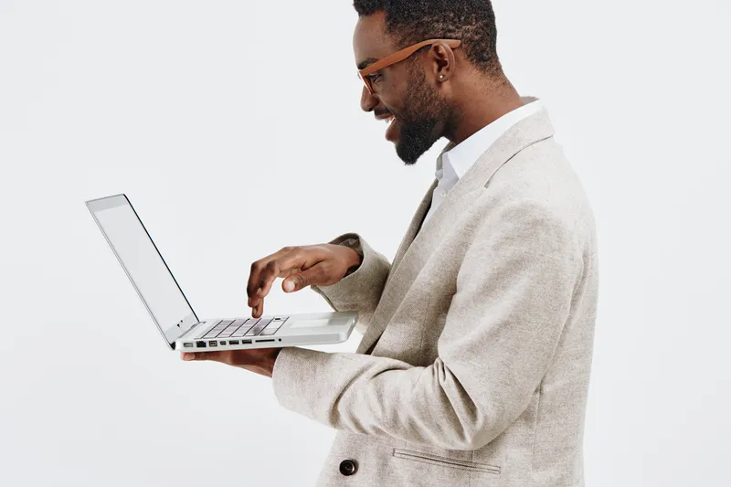 Professional young man using a laptop computer, wearing glasses and a beige blazer, standing against a plain white background. Male office worker or freelancer, technology, work, modern lifestyle, cor