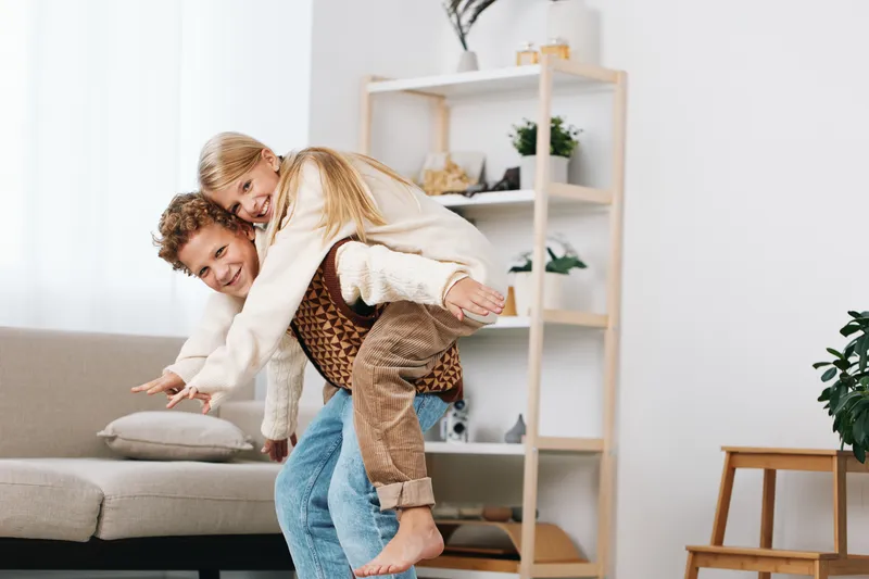Joyful couple share a playful piggyback ride indoors, smiling as they lift each other in a light-filled living room with a sofa, shelves, and a touch of greenery.