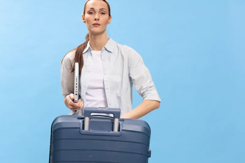 Confident young woman with long hair holding suitcase and laptop stands against blue background. Travel and business concept with casual outfit and determined expression in studio lighting.