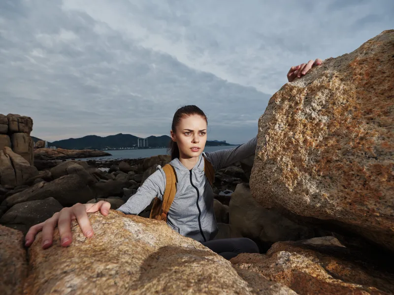 Young female hiker navigating rocky terrain with determination, wearing a grey jacket and backpack, showcasing a cloudy day atmosphere and adventurous spirit