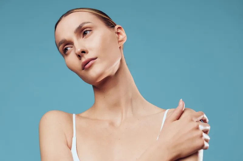 Young woman with clear skin looks away thoughtfully while touching her shoulder isolated on blue background, beauty portrait with natural makeup and calm expression.