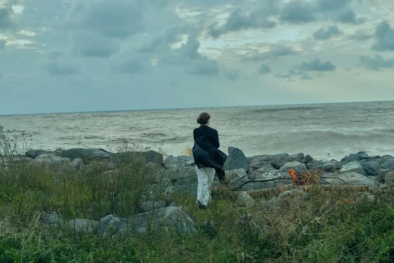 Figure in dark jacket standing on rocky shoreline, looking toward the sea under a cloudy sky, a calm moment of solitude by the coast.