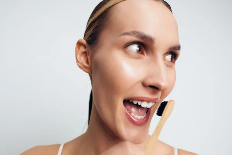 Young woman smiling with open mouth holding a toothbrush near her face, bright skin, natural makeup, white background, dental hygiene concept, cheerful expression