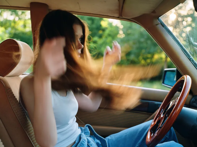 Young woman enjoying music while driving a classic car, with flowing hair and a carefree expression on her face in a vibrant green outdoor setting.