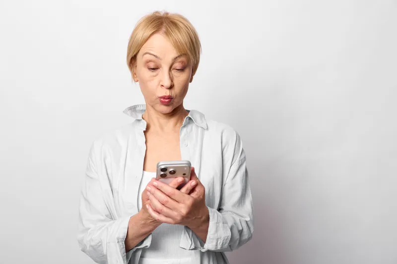Stressed woman holds a smartphone, with anxious worried expression on a plain white background. Concept of anxiety, mental health, communication, and technology for negative news.