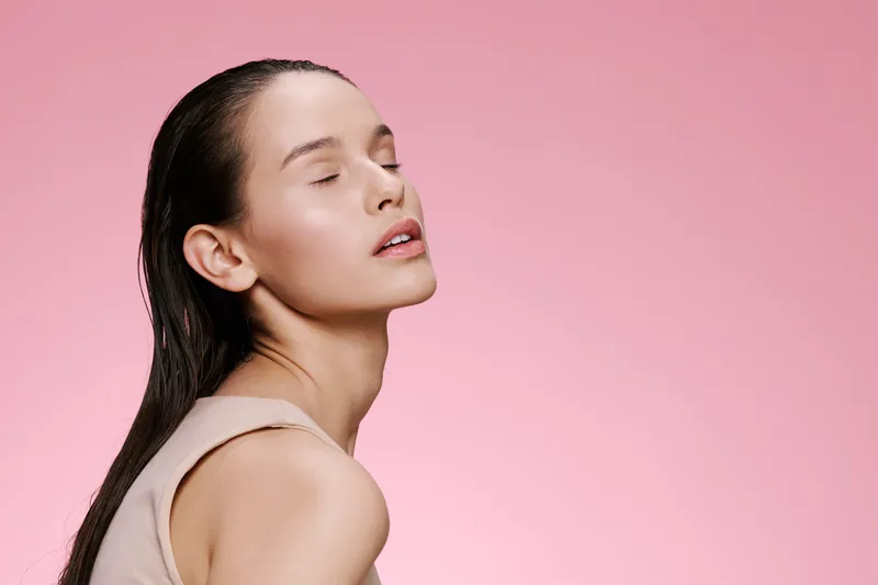 Elegant young woman with wet hair and closed eyes, showcasing natural beauty against a soft pink background, ideal for health and beauty concepts