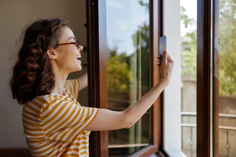 Woman lifestyle home, smiling woman takes selfie by open window, bright living room, casual pose, natural light, cozy interior, everyday moment, modern decor, green outdoor trees visible
