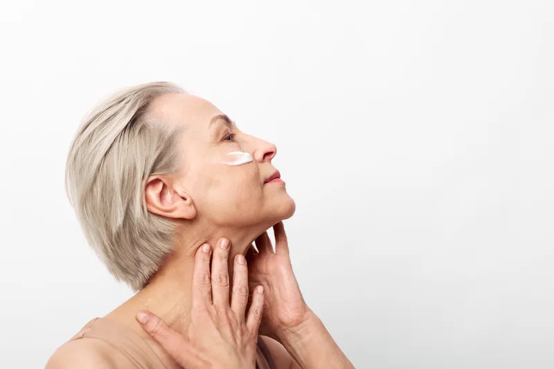 Mature woman with short gray hair applies white face cream on cheek, touching neck gently. Skincare and self-care concept, natural beauty and aging gracefully on clean white background.