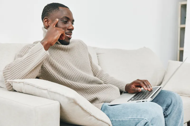 Comfortable African man working on a laptop while sitting on a couch in a cozy living room, wearing a casual sweater, showcasing productivity and relaxation