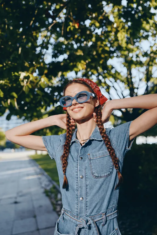 smiling woman with braids wearing denim jumpsuit and sunglasses outdoors in sunny park with green trees background