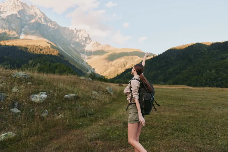 A young woman with long brown hair wearing a beige shirt and green shorts hikes in a mountainous landscape while pointing towards the distant peaks under a clear sky.