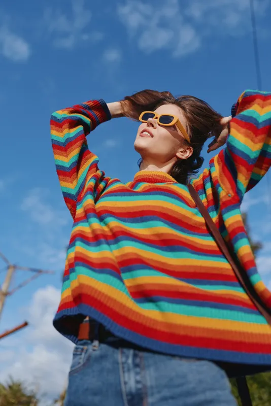 Young woman in a colorful striped sweater and denim jeans wears yellow sunglasses, smiling and leaning back against blue sky outdoors. Casual outfit, confident travel and lifestyle portrait.