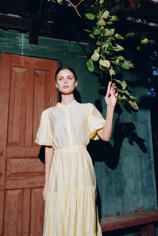 woman wearing a flowing yellow dress stands by a doorway amid greenery, creating a fashion forward portrait with elegant calm and a vibrant natural backdrop.