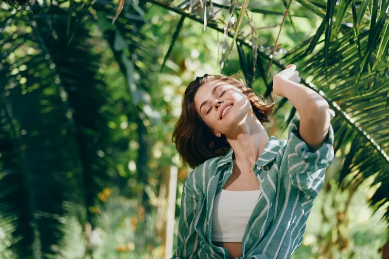 A young woman enjoys a sunny outdoor moment among lush greenery, smiling with eyes closed, wearing a striped shirt over a white top, bathed in warm light.