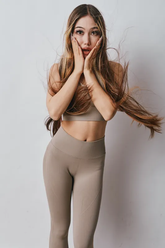 Young woman with long hair wearing beige activewear posing with surprised expression and hands on face against plain background for fitness or lifestyle concept.