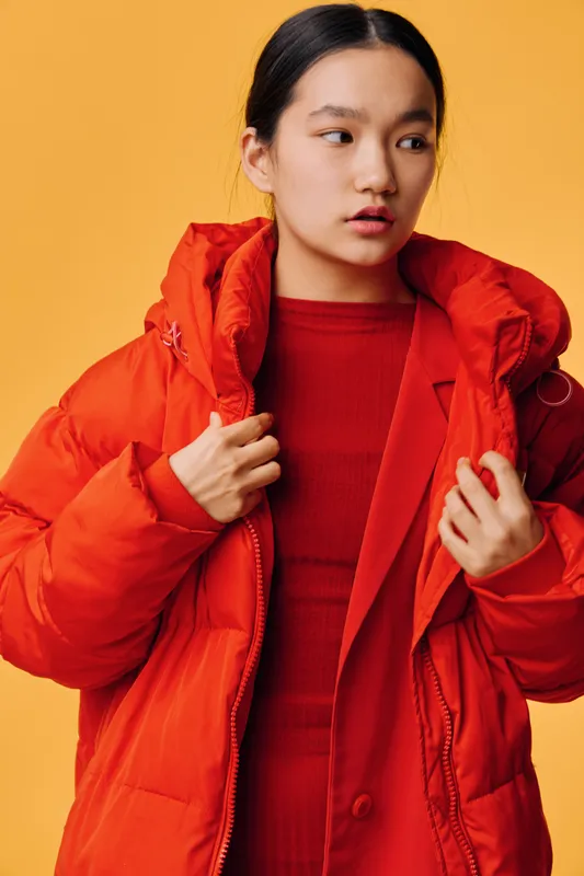 Young woman wearing bright red puffer jacket and red sweater standing against orange background with thoughtful expression, casual winter fashion and warm clothing style.