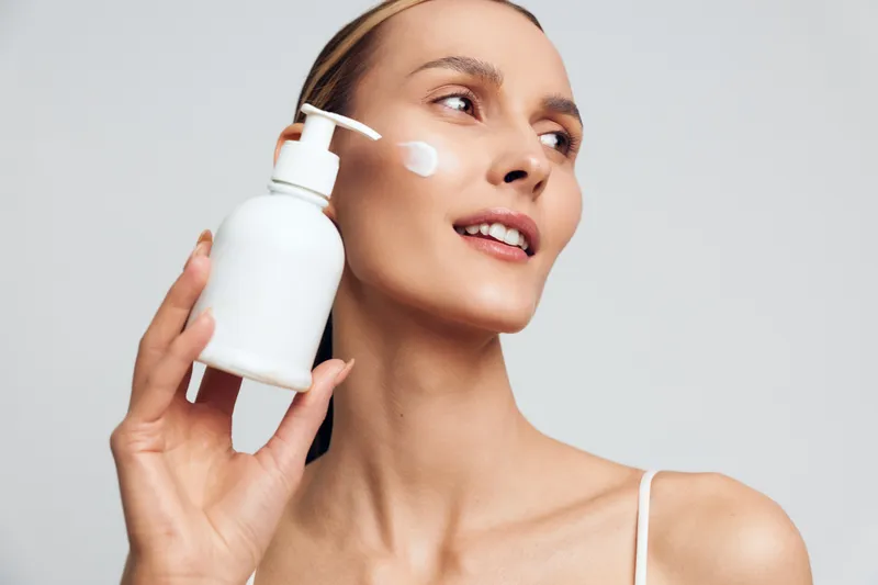 Woman face skincare serum lotion cosmetics beauty wellness wellbeing spa concept, young woman holds pump bottle and applies white cream on cheek, closeup in soft studio lighting on clean background
