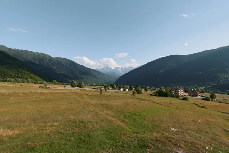 A tranquil countryside landscape unfolds across a wide valley framed by gentle hills. Sparse trees dot the fields beneath a clear blue sky, evoking peaceful, timeless natural beauty.