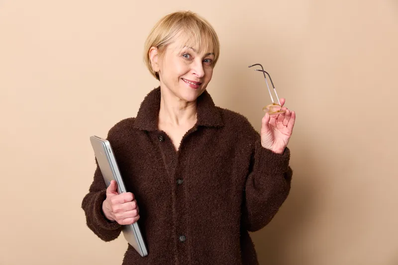 Woman senior smartphone glasses holding smile indoors studio beige portrait, relaxed female with short hair in dark coat holding tablet and eyeglasses on plain background