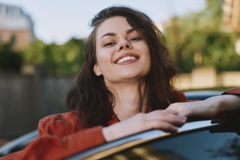 A smiling woman leans on a sunlit car outdoors, showcasing casual lifestyle and carefree confidence. Bright urban setting, warm tones, relaxed pose, everyday happiness in frame