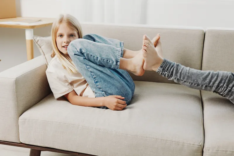 A playful young girl with blonde hair lies on a beige sofa wearing casual jeans and a white shirt, engaging in a fun moment with a person extending feet toward her. Concept of childhood, play, and