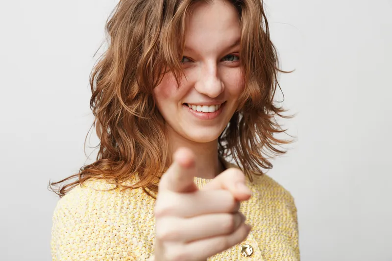Smiling young woman with wavy brown hair points finger at camera, wearing yellow sweater. Friendly confident gesture on light background. Concept of communication and positivity.