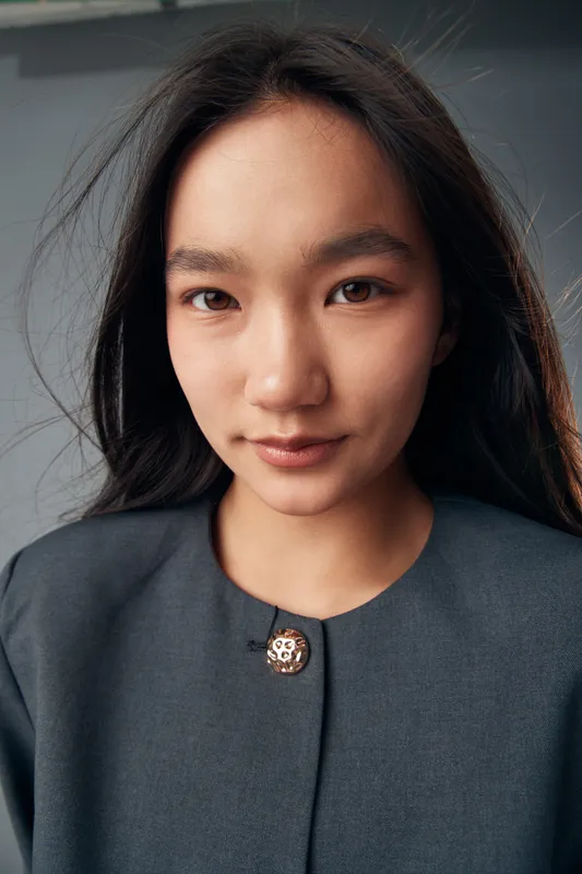 Portrait of Asian woman with long hair looking at camera with calm expression wearing dark gray jacket with decorative button on light background.