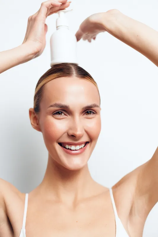 Smiling young woman holds white cosmetic bottle above head with both hands shows skincare product application concept isolated on white background with positive facial expression.