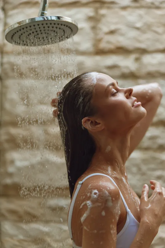 woman showering indoors under rainfall showerhead with wet hair, relaxed expression, beige stone tile background, intimate moment with water droplets