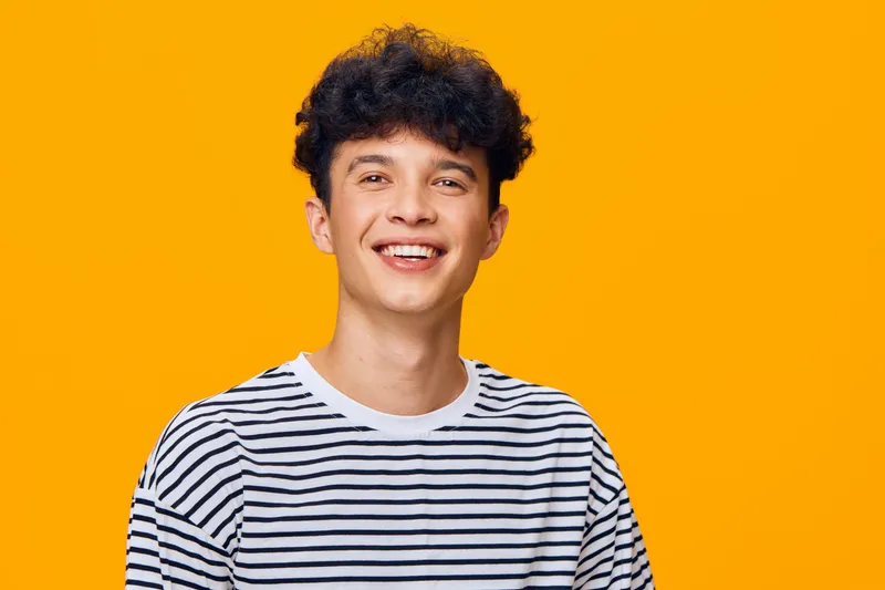 Smiling man with curly hair wearing a striped shirt stands against bright orange background, conveying happiness and youthful energy in vibrant studio lighting.