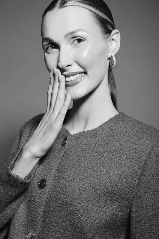 Young white woman with light skin and blonde hair smiles shyly, hand near face, wearing textured jacket on grey background.