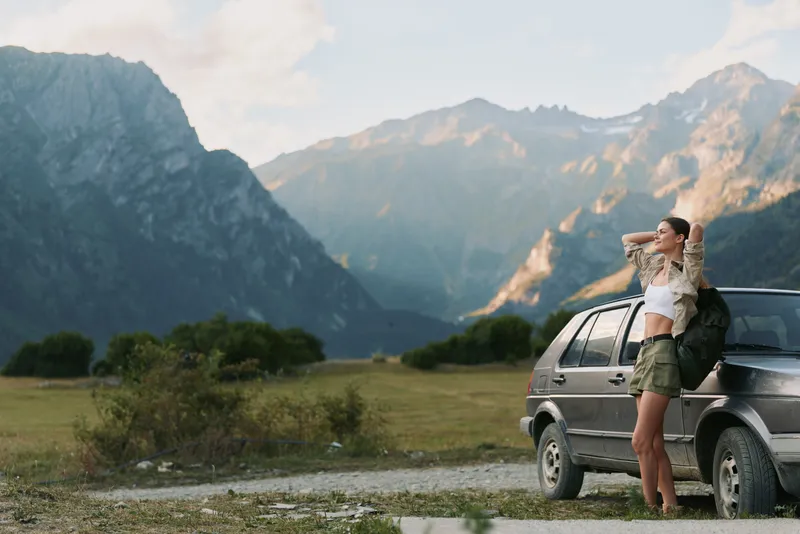 One young woman stands beside a silver car in a mountain valley, sunlit and relaxed, enjoying the moment.
