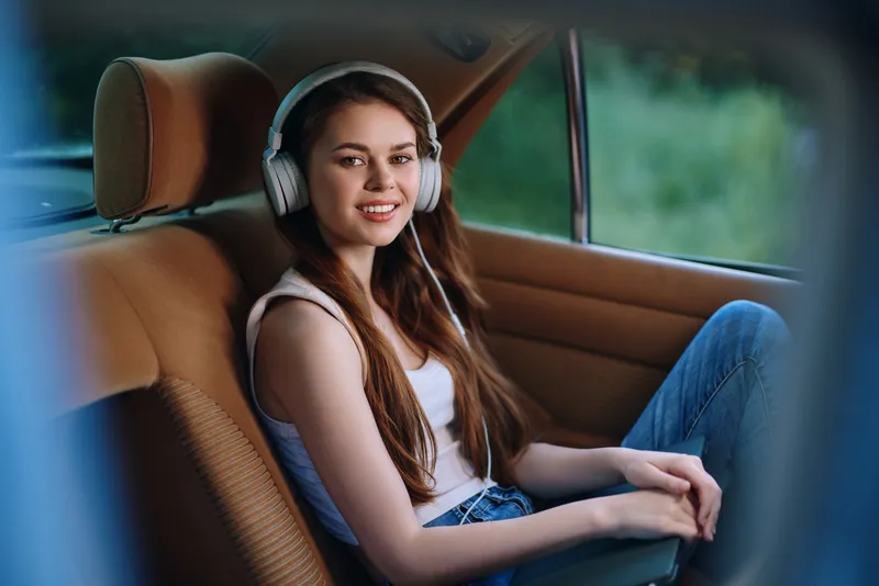 Young woman enjoying music in a car with headphones, casual outfit, natural smile, green background, lifestyle concept, relaxation, freedom.