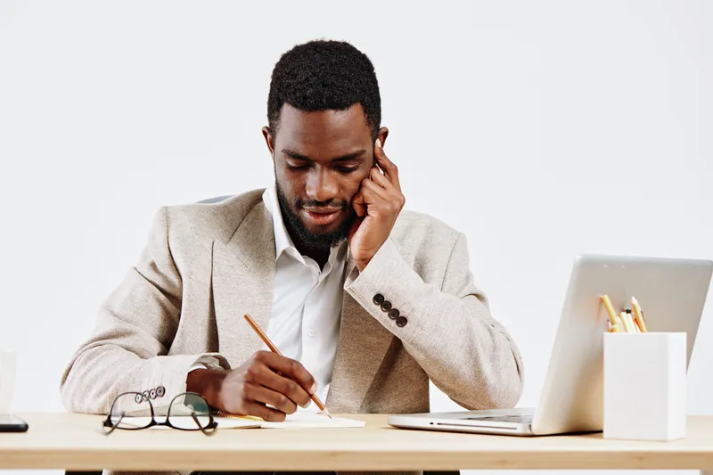 Professional man working at desk with laptop and notebook, wearing beige blazer and white shirt, focused on writing, modern office environment, casual business style, clean background, concept of prod