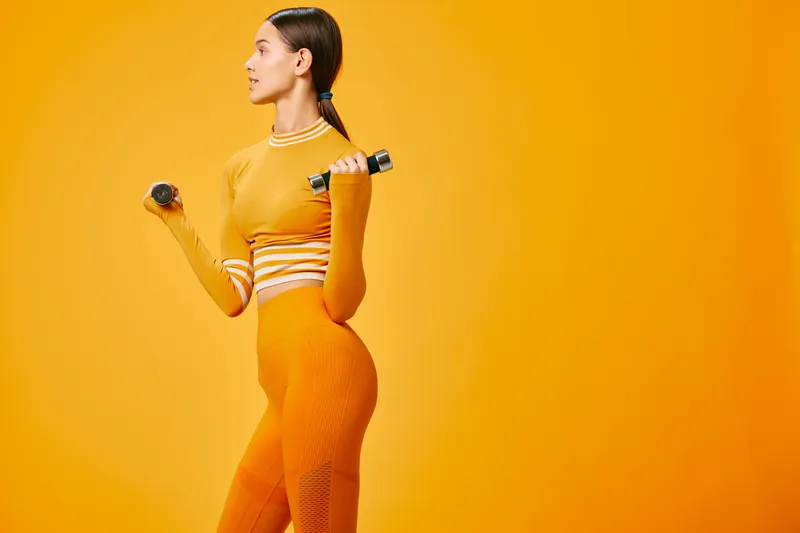 Woman fitness, sport portrait in a bold orange studio with dumbbells, capturing strength, balance and motivation during a focused workout session with dynamic pose and athletic energy.